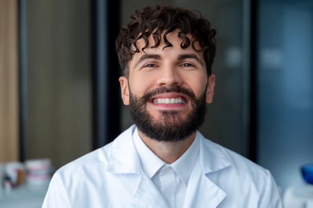 A confident, bearded man in a white lab coat smiles, possibly due to clear braces at Carpinello Orthodontics in Edgmont, PA.
