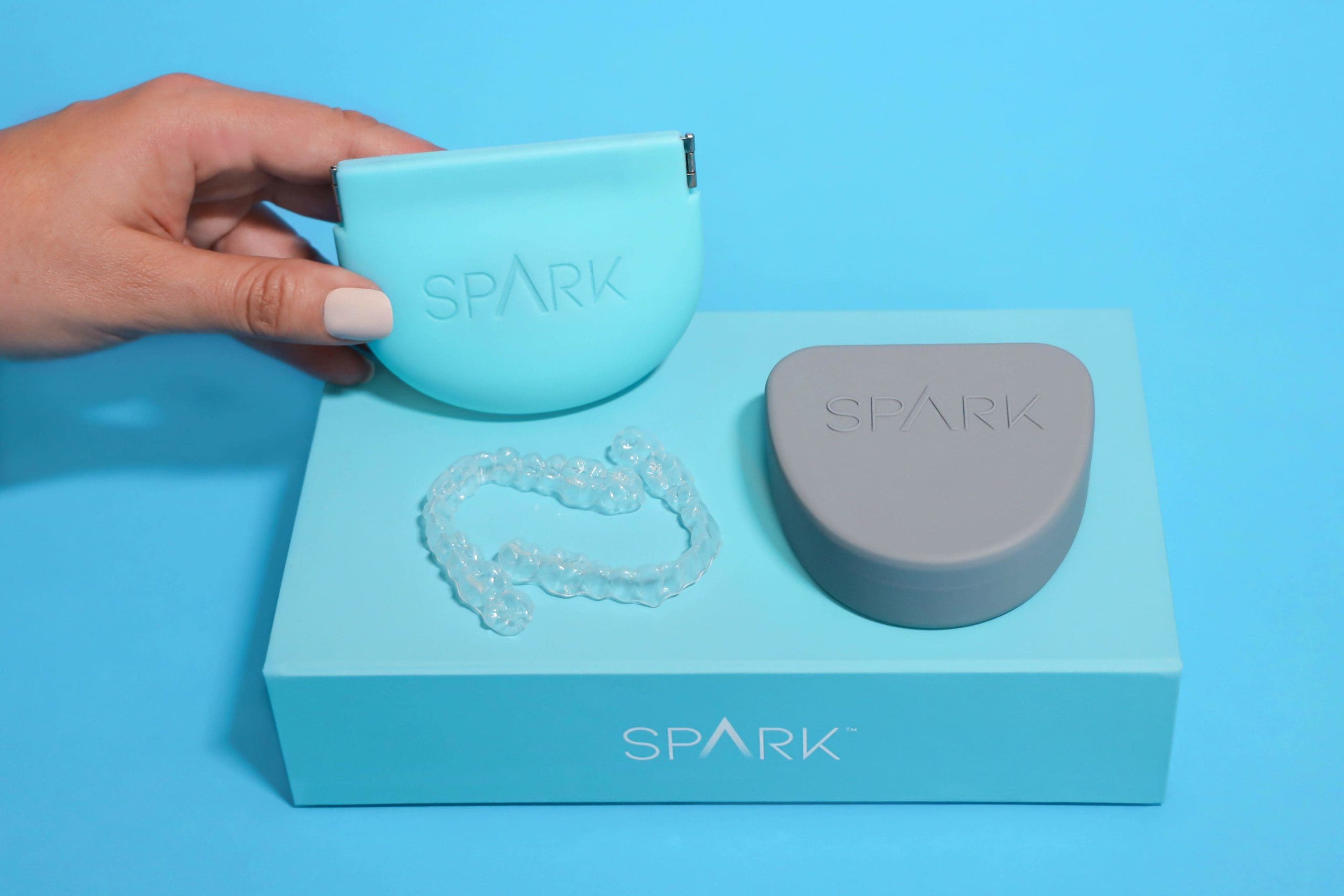 At Carpinello Orthodontics in Edgmont PA, a hand holds a light blue SPARK aligner case above a box with a gray case and clear aligner on a blue background.